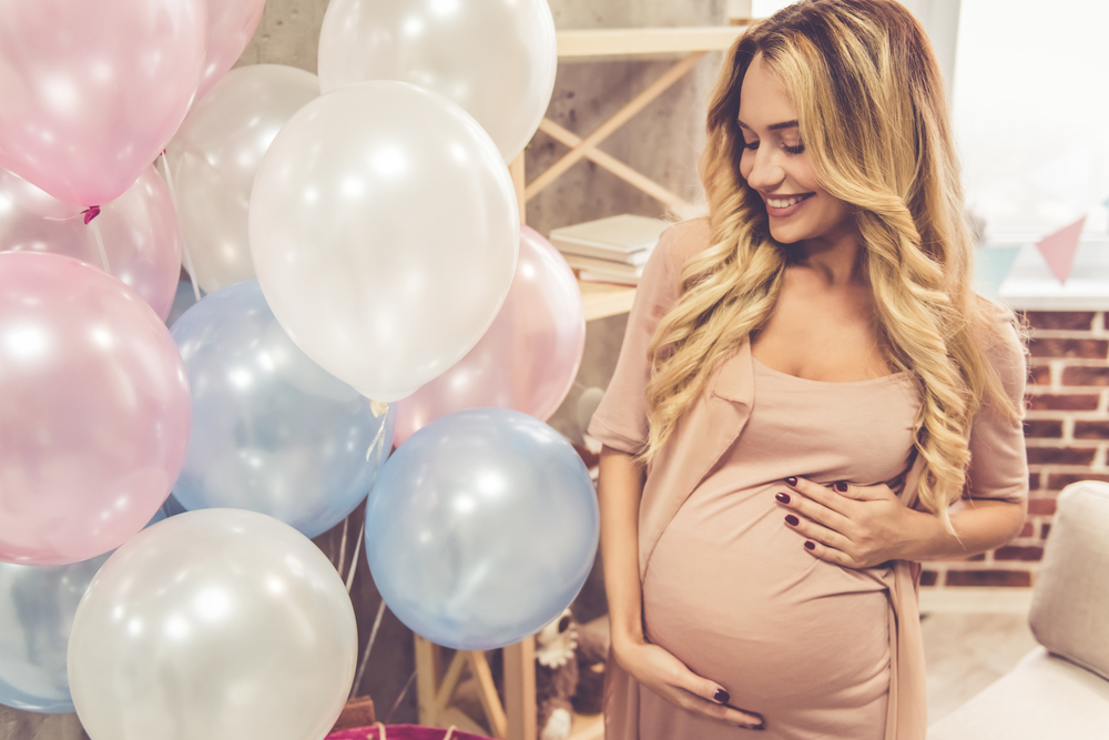 A pregnant lady holding her belly, stood next to some balloons. 